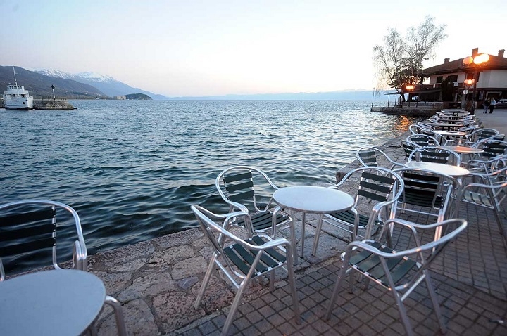 Поглед кон Охридско Езеро од едно кафуле, кое има поставено столчиња и маси на брегот на езерото.
